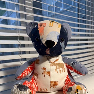 K&ouml;nnte beinhalten: Ein handgefertigter Patchwork-Teddyb&auml;r mit schwarzer Nase und Knopfaugen. Der Kopf und die Ohren des B&auml;ren sind marineblau mit einem Muster aus Tieren und Sternen. Der K&ouml;rper weist verschiedene Stoffmuster auf, darunter rote, wei&szlig;e und Tiermuster. Der Name "Patrick" ist auf einem Pfoten sichtbar.