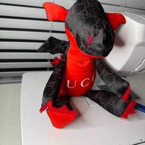 K&ouml;nnte beinhalten: Ein Pl&uuml;schdrachen-Spielzeug mit rotem K&ouml;rper und schwarzen Fl&uuml;geln und Kopf. Der schwarze Stoff hat ein sich wiederholendes Muster. Das Wort "UC" ist auf die Brust des Drachen gedruckt. Der Drache sitzt auf einer wei&szlig;en Oberfl&auml;che.