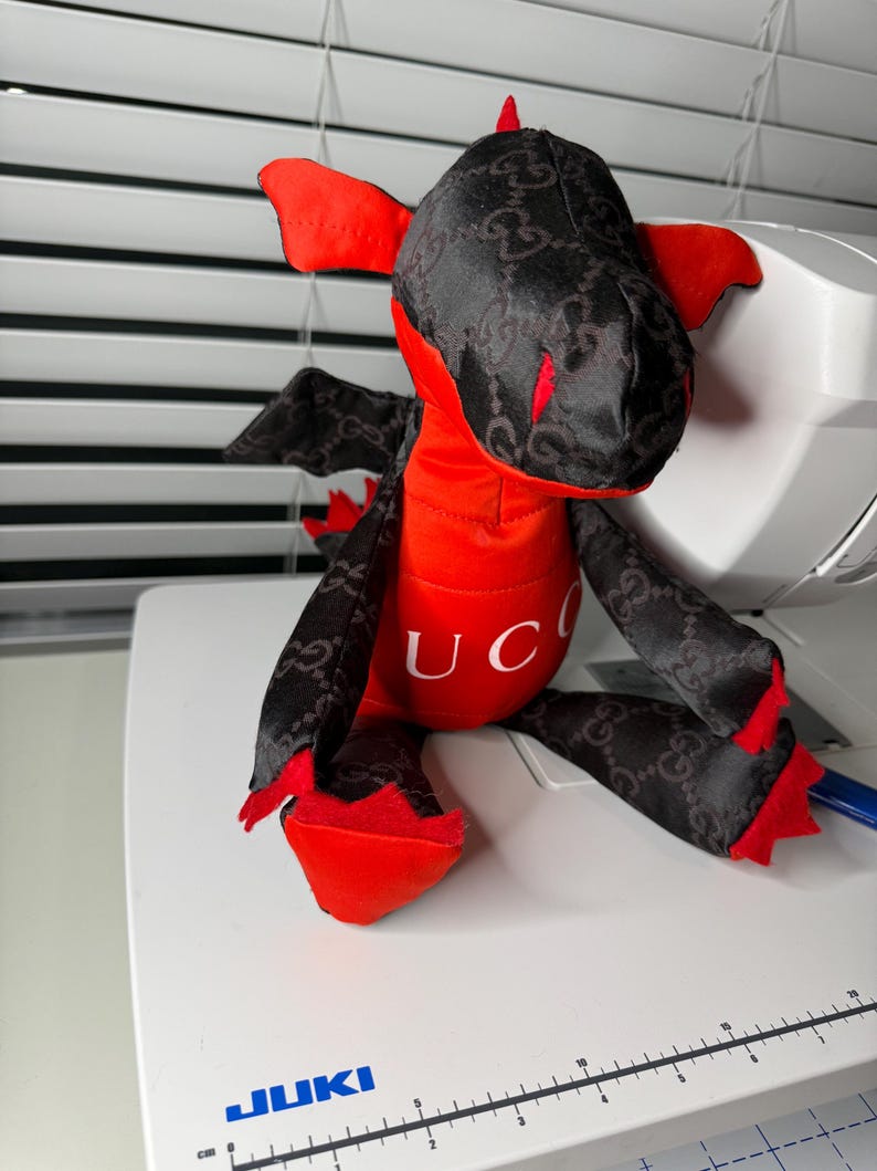 K&ouml;nnte beinhalten: Ein Pl&uuml;schdrachen-Spielzeug mit rotem K&ouml;rper und schwarzen Fl&uuml;geln und Kopf. Der schwarze Stoff hat ein sich wiederholendes Muster. Das Wort "UC" ist auf die Brust des Drachen gedruckt. Der Drache sitzt auf einer wei&szlig;en Oberfl&auml;che.