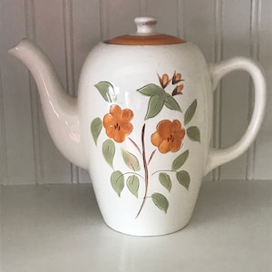 Puede incluir: Tetera de cerámica blanca con tapa marrón y diseño floral con flores naranjas y hojas verdes.