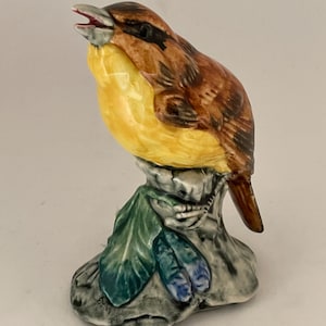 Puede incluir: Una figurita de cerámica de un pájaro amarillo y marrón posado en una roca con hojas verdes y azules. El pájaro tiene un pico largo y está cantando.