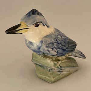 Puede incluir: Una figurita de cerámica azul y blanca de un pájaro posado en una roca verde. El pájaro tiene un pico largo y puntiagudo y un cuerpo detallado y texturizado.