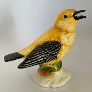 Puede incluir: Una figurita de cerámica de un pájaro amarillo con alas negras y una base verde. El pájaro tiene un pico amarillo y ojos negros.