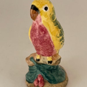 May include: A small ceramic figurine of a yellow and green parakeet perched on a branch with red berries. The bird has a pink breast and a brown beak.
