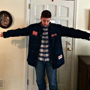 May include: A blue denim jacket with red and white patches. The patches have the words "Richmond" and "Richmond Oil" on them. The jacket is worn over a plaid shirt and blue jeans.