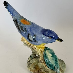 Puede incluir: Una figurita de pájaro de cerámica azul y amarilla posada en una rama con hojas verdes. El pájaro tiene una cola larga y un pico pequeño.