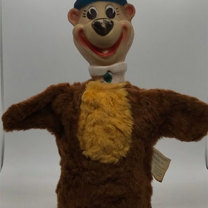 May include: A brown and yellow plush puppet with a blue hat. The puppet has a smiling face with black eyes and a black nose. The puppet is a character from the cartoon show "Yogi Bear".