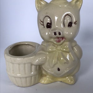 May include: A ceramic planter shaped like a pig wearing a bow tie. The planter has a separate pot for planting. The pig is light yellow with brown eyes and a pink tongue.