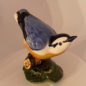 Puede incluir: Una figurita de cerámica de un pájaro azul posado en una rama. El pájaro tiene marcas negras y blancas en la cabeza y una barriga amarilla. La rama es marrón y la base es verde.