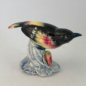 May include: A ceramic bird figurine with black, yellow, red, and white painted details. The bird is perched on a blue and white ceramic base with a pink flower.