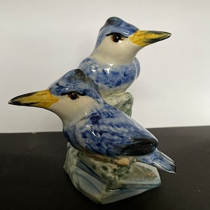 Puede incluir: Dos pájaros de cerámica azul y blanco posados ​​en una roca. Los pájaros tienen picos amarillos y ojos negros. La roca está pintada en tonos de azul, verde y marrón.