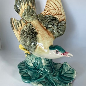 May include: A ceramic bird figurine with green, brown, and yellow painted details. The bird is perched on a green leaf with a white background.