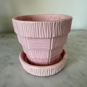 Op de afbeelding: Een roze keramische bloempot met bijpassende schotel. De pot heeft een gestructureerd, gelaagd ontwerp met verticale en horizontale lijnen. De kleur is een zacht, gedempt roze en de pot lijkt van keramiek te zijn.