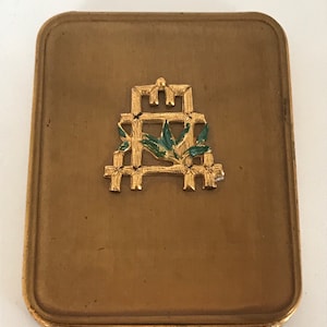 May include: A gold metal compact with a decorative design of a stylized bamboo plant with green leaves.