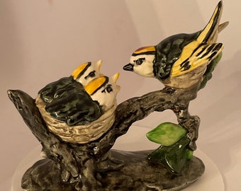 Może przedstawiać: Ceramiczna figurka ptaka z żółtymi i czarnymi oznaczeniami, siedzącego na gałęzi i patrzącego w dół na trzy pisklęta w gnieździe. Gniazdo jest zrobione z plecionych gałązek, a gałąź jest brązowa i fakturowana.