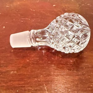 May include: A clear glass decanter stopper featuring a spherical top with a diamond-cut geometric pattern and a smooth, frosted tapered base designed to fit securely into the neck of a glass bottle or decanter vessel.