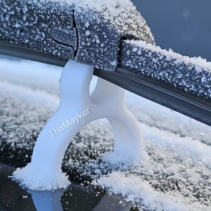 May include: A close-up shot of a car windscreen wiper blade heavily coated in snow and ice. A white, Y-shaped support is holding the wiper blade up. The text "ThaMayker" is visible on the support, preventing the wiper from freezing to the glass.