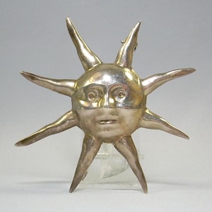 May include: A silver sun pendant with a face and eight rays. The pendant is a stylized sun with a human face.