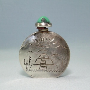 May include: A small, silver-coloured perfume bottle with a green stone stopper. The bottle features an etched design of a pyramid, sun, and mountains. A cactus is also etched on the bottle. The bottle is round and has a small, red band near the stopper.