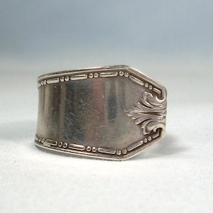 Może przedstawiać: Srebrny pierścionek wykonany z łyżki, z prostokątną powierzchnią i dekoracyjnymi krawędziami z koralików oraz motywem kwiatowym. Pierścionek ma wygląd vintage, prezentujący wzór ze srebra.