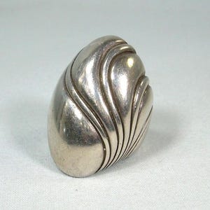 May include: A silver-toned, oval-shaped ring with a textured, wave-like design. The ring has a smooth, polished surface and a detailed pattern of curved lines. The design is reminiscent of a seashell or a stylized leaf.