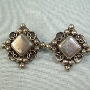 Könnte beinhalten: Ein Paar silberfarbene Metallbroschen mit quadratischem Design. Jede Brosche hat ein erhabenes Quadrat in der Mitte und ist von einem dekorativen Rand aus kleinen Perlen und Schnörkeln umgeben.