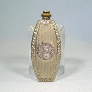 Op de afbeelding: Een vintage, ovaalvormige parfumfles met een goudkleurige metalen afwerking. De fles heeft een centraal embleem met een berggeit en de tekst "Rocky Mtn Nat'l Park". Decoratieve bladeren omlijsten de randen.