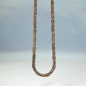 May include: A metallic rose gold-toned chain necklace featuring a dense, intricate woven wheat or foxtail link pattern. The chain hangs in a U-shape, displaying a consistent, textured, and braided appearance throughout its entire length.