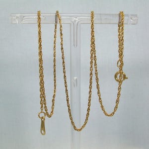 May include: A gold-colored necklace with a rope chain design. The necklace is displayed on a clear stand, showcasing its delicate links and clasp. The chain is approximately 20 inches long.
