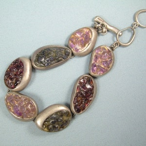 Könnte beinhalten: Ein silbernes Armband mit einem Schiebeverschluss verfügt über sieben ovale Steine mit einem mosaikartigen Muster in Violett-, Rosa- und Grüntönen.