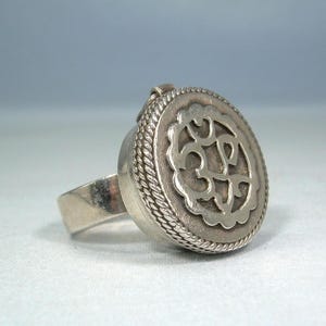 Könnte beinhalten: Ein silberfarbener Ring mit einer großen, kreisförmigen, dosenartigen Fassung. Die Oberseite der runden Fläche zeigt ein komplexes, ausgeschnittenes Om-Symbol, umgeben von einem gewellten Rand und einer dekorativen Kordelverzierung am Gehäuse.