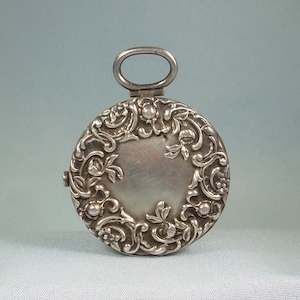 Puede incluir: Un medallón de plata con un diseño floral. El medallón tiene un bucle en la parte superior para sujetar una cadena.