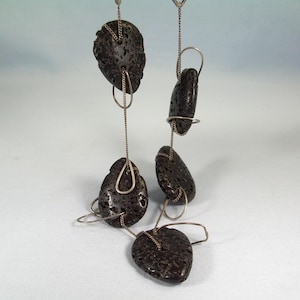 以下が含まれることがあります： 繊細なシルバーチェーンとフープで吊るされた、暗く質感のある石が特徴的な手作りのイヤリング。 イヤリングは有機的で自由なデザインで、それぞれの石がユニークな形をしています。 全体的な美しさはモダンでアースカラーです。