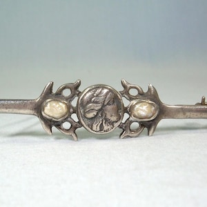 Puede incluir: Un broche de barra plateado con un diseño antiguo. El broche presenta un medallón central con una imagen de perfil, flanqueado por dos piedras ovaladas de color blanquecino. Los extremos del broche tienen formas decorativas y estilizadas.