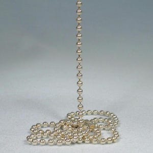 Puede incluir: Un collar de cadena de bolas de color plateado, con una hebra vertical de esferas conectadas que se extiende desde una parte enredada en la base. La cadena se muestra sobre un fondo gris claro.