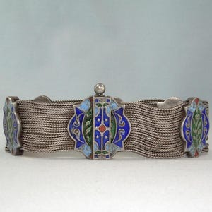 May include: Silver-toned bracelet with intricate woven chain links. The bracelet features decorative enamel panels in blue, green, and red. The central panel has a small, round silver accent.