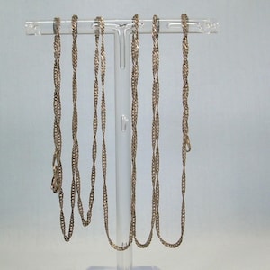 May include: Several silver-toned chain necklaces with a twisted rope design are displayed on a clear plastic jewelry stand. Each chain has a clasp at the end. The necklaces hang vertically, showcasing their intricate woven pattern.
