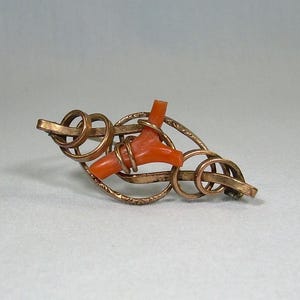 Puede incluir: Un broche dorado con una pieza de coral en el centro. El broche está hecho de círculos de metal entrelazados.