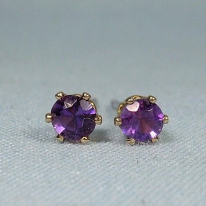 May include: A pair of stud earrings featuring round, faceted purple gemstones set in gold-colored metal. The earrings are displayed on a light blue surface, showcasing their vibrant color and simple design.