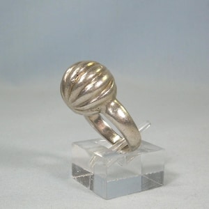 May include: A silver ring with a ribbed, spherical design. The ring is displayed on a clear, square stand against a light blue background. The ring's band is smooth and rounded, and the spherical top has a textured, grooved pattern.