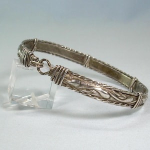 May include: Silver-toned bracelet with a decorative design. The bracelet features a woven pattern and a hook clasp. The design includes a detailed floral pattern on the front. The bracelet is displayed on a clear stand.