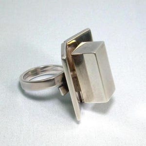 May include: A silver ring with a rectangular design. The ring features a thick band and a large, geometric centerpiece. The centerpiece has a boxy shape with a flat, rectangular face. The ring is set against a white background.