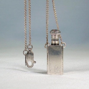 May include: A silver perfume bottle pendant necklace. The rectangular bottle has a screw-on cap and is suspended from a delicate chain. The clasp is visible. The background is a light blue.