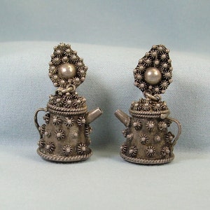 May include: A pair of silver teapot-shaped dangle earrings with a floral design. The earrings are made of silver and have a detailed floral pattern.