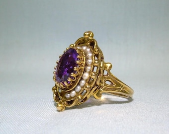 May include: A gold ring with a large purple gemstone surrounded by small white pearls. The ring has a delicate, intricate design.