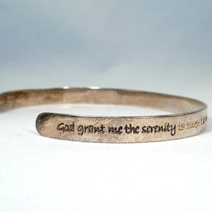 Könnte beinhalten: Ein silberfarbenes Armreif-Armband mit dem eingravierten Text "Gott gib mir die Gelassenheit, die Dinge zu akzeptieren, die ich nicht ändern kann." Das offene Armband wird auf einer weißen Oberfläche gezeigt.