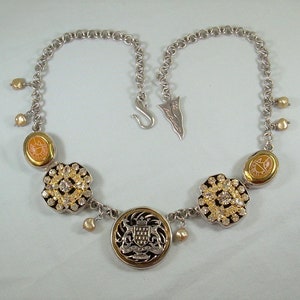 May include: A silver chain necklace with a gold pendant featuring a coat of arms design. The necklace also has several smaller pendants with rhinestones and orange enamel.