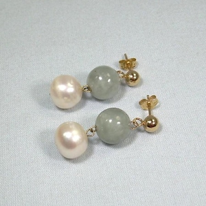 May include: A pair of drop earrings with a light pink pearl and a grey-green bead. The earrings have gold-coloured posts and connecting wires. The jewellery is displayed on a light blue surface.