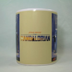 May include: White ceramic mug with a beige background and a blue and purple image of a character from the Star Wars: The Mandalorian TV series. The text "STAR WARS THE MANDALORIAN" is printed in black on the mug.