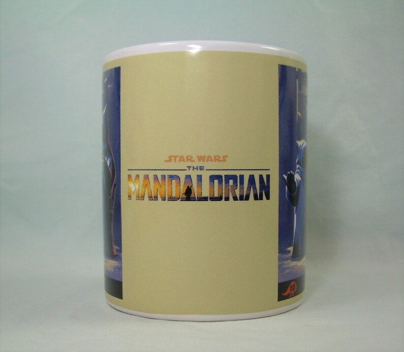 May include: White ceramic mug with a beige background and a blue and purple image of a character from the Star Wars: The Mandalorian TV series. The text "STAR WARS THE MANDALORIAN" is printed in black on the mug.